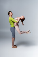 strong ballet dancer lifting ballerina in bodysuit on grey