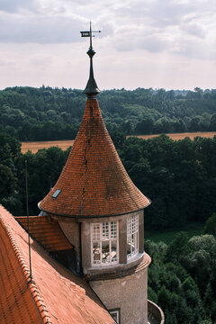 Castle tower with gallery and a red pointed roof with wind rose on top. Wicker chairs and a table behind the open white windows of the castle tower, field and forest in the background.