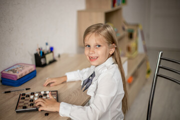 A boy in a white shirt plays checkers while sitting at a desk.