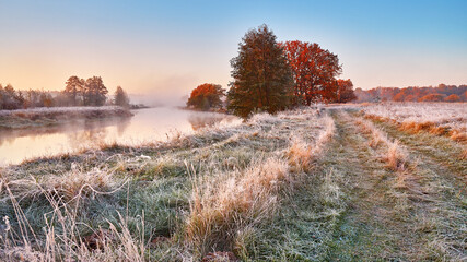 Foggy river and meadow fall sunrise. Grass covered with white frost on riverbank in early morning panorama. Dirt road on field, oak tree with orange leaves