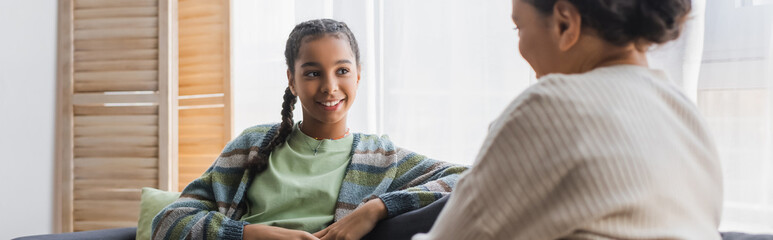 smiling african american girl looking at blurred mother during conversation at home, banner