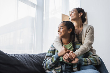 happy african american mother and daughter sitting on couch and looking through window at home