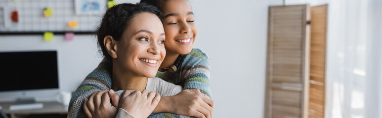 teenage african american girl with closed eyes embracing happy mom at home, banner