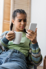 discouraged african american girl with cup of tea frowning while looking at smartphone, blurred background