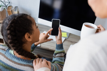 african american girl using cellphone with blank screen near computer monitor and blurred mom with...