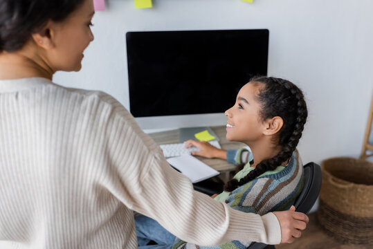 Happy African American Girl Looking At Blurred Mother While Doing Homework Near Monitor With Blank Screen