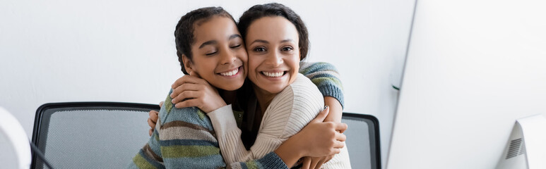happy african american woman and teenage girl embracing near computer monitor, banner