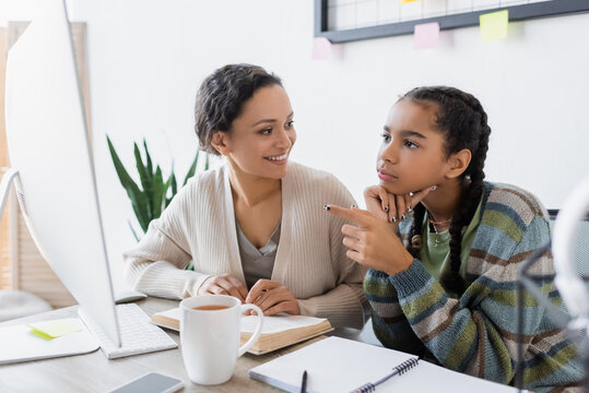 african american girl pointing at computer monitor while doing homework with smiling mother