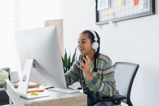 excited african american teenage girl in headphones gesturing while looking at monitor