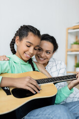 african american teenage girl playing acoustic guitar near happy mom at home