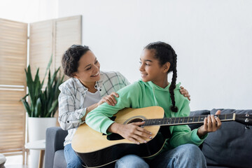 smiling african american girl looking at happy mom while playing guitar at home
