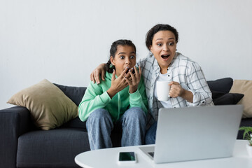 shocked african american woman with teenage daughter watching movie on blurred laptop