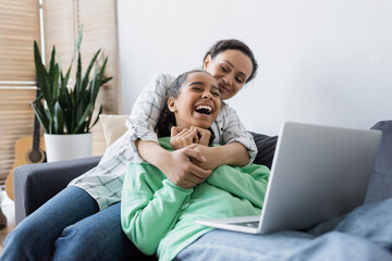 african american woman hugging laughing daughter while watching film on laptop