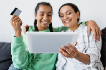 blurred african american woman holding digital tablet near happy daughter with credit card