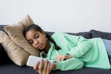 upset african american teen girl using mobile phone while lying on sofa