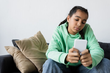 frustrated african american teen girl messaging on blurred mobile phone while sitting on couch