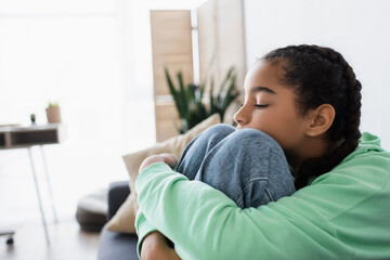 upset african american teenage girl hugging knees while sitting with closed eyes at home