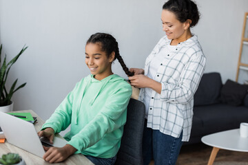smiling african american woman braiding hair of teenage daughter using laptop while studying at home