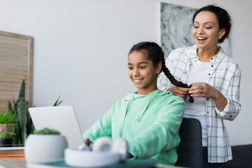 cheerful african american woman braiding hair of daughter doing homework on laptop