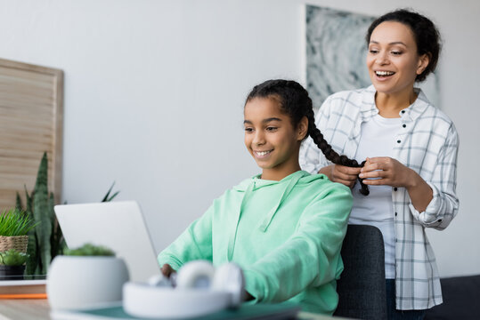 cheerful african american woman braiding hair of daughter doing homework on laptop