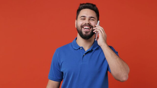Happy calm smiling young bearded brunet man 20s years old wears blue t-shirt hold use talk on mobile cell phone conducting pleasant conversation isolated on plain red orange background studio portrait