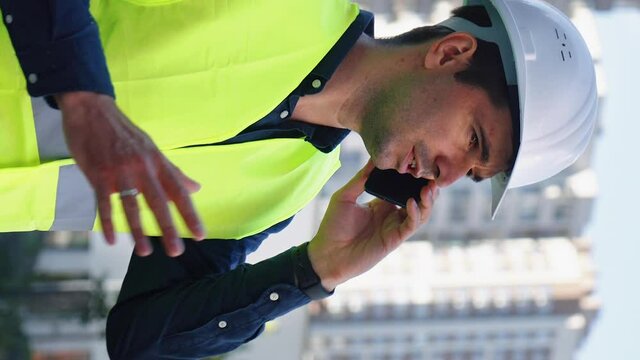 Vertical Screen: Engineer wearing hard hat and safety vest talking on phone while walking against multistory buildings. Building contractor discussing plans with architect. Concept of communication