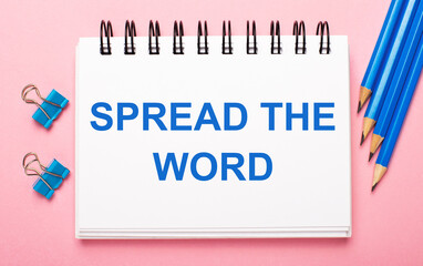 On a light pink background, light blue pencils, paper clips and a white notebook with the text SPREAD THE WORD