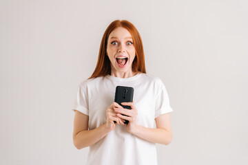 Fototapeta premium Studio portrait of happy excited young woman open mouth reading good online message using mobile phone on white isolated background. Cheerful lady holding smartphone in hands and looking at camera.