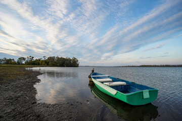           A small green boat is moored by the lake. The boat is empty, no one is in it. The water reflects the sky and the forest in the background.
