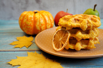 Belgian gluten-free pumpkin waffles lie on a brown plate with a dry slice of lemon, pear, apple and anise stars on a blue wooden table. side view. maple leaves