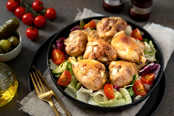 Fried chicken legs with salad in a plate on a dark kitchen table. Cooked poultry meat, pickles and tomatoes on the culinary background of closeup