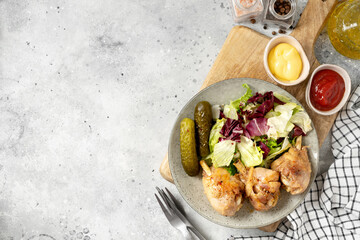 Fried chicken legs with salad and pickles in a plate on a light gray kitchen table. Cooked poultry meat on a culinary background top view with a copyspace
