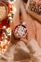 cup of cocoa or coffee drink with marshmallows in female holding hands over the legs of woman in knitted sweater relaxing and enjoyment the holidays in cozy bed at home on christmas or new year eve