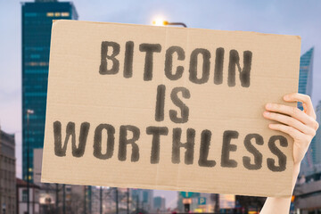 The phrase " Bitcoin is worthless " on a banner in men's hand with blurred background. Bankruptcy. Economic. Crisis. Savings. Risky. reduction. Fraud. Technology. Volatility