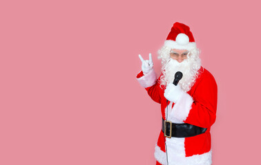 Stylish adult male vocalist, dressed as Santa Claus, shouts into the microphone, with a sign from the fingers of Victoria, victory, peace, forever. Isolate on a pink background