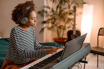 Multiracial female musician sitting on bed and playing melody on electronic musical instrument