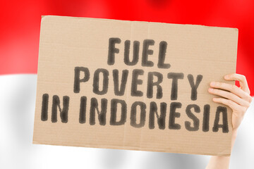 The phrase " Fuel poverty in Indonesia " on a banner in men's hand with blurred Indonesian flag on the background. Economic. Expensive. Depressed. Gasoline. Fossil. Problem. Supply. Social issues