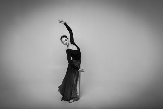Black and white photo, beautiful, pretty ballerina showing various exercises, looking straight into the camera in a long dress on a light background. Selective focus, movement, low key