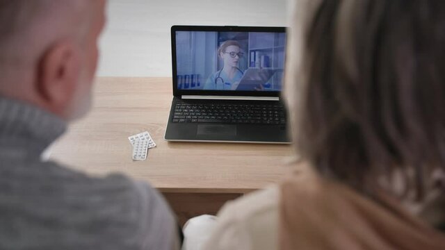 Old Patients Consult Doctor Online Via Video Communication Using Modern Technologies, Elderly Man And Woman Sitting At Table, Over Shoulder Laptop View