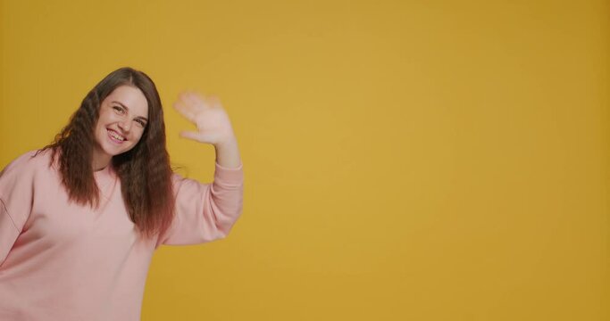 Smiling young girl blogger greeting subscribers wave hand say hello on yellow background with copy space for advertising