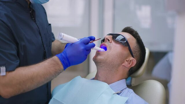 Caucasian brunette man in dentist chair with unrecognizable doctor in gloves using ultraviolet curing light in slow motion. Male patient and orthodontist in hospital indoors. Slow motion