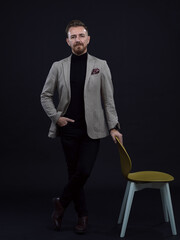 Portrait of adult businessman wearing trendy suit and sitting in modern studio on stylish chair against the black background