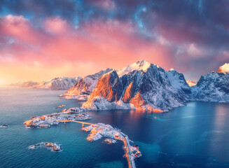 Beautiful landscape with blue sea, snowy mountains, rocks and islands, village, rorbu, road, bridge and pink sky at sunrise. Aerial view. Hamnoy in snow in winter in Lofoten islands, Norway. Top view - obrazy, fototapety, plakaty