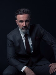 Portrait of adult businessman wearing trendy suit and sitting in modern studio on stylish chair against the black background