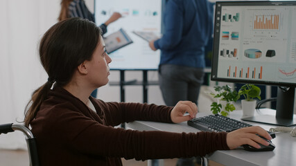 Invalid handicapped disabled financial expert sitting in wheelchair working in busy start up company with young team, analysing economic statistics, checking graphs for new project