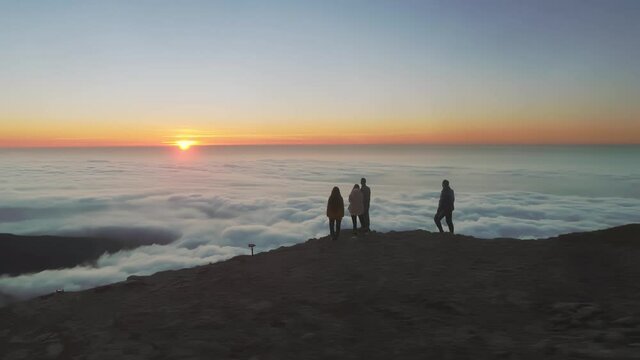 Aerial flight by drone around the group of peoples silhouettes at mountain top at sunrise against clouds and sun