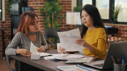 Business partners analyzing statistics on papers in startup office. Asian women working together to develop financial strategy, planning marketing project with documents. Work productivity