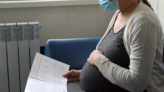Young Pregnant Woman In Medicine Protective Mask Reading On Couch Waiting For Appointment Medical Control On Hospital. Visit Gynecologist Doctor At Clinic. Examine Expectant Belly Healthcare Check Up