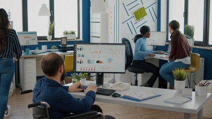 Paralized employee talking with african team leader woman sharing information pointing on graphs from computer. Colleague giving feedback discussing solution for project deadline in strat up office