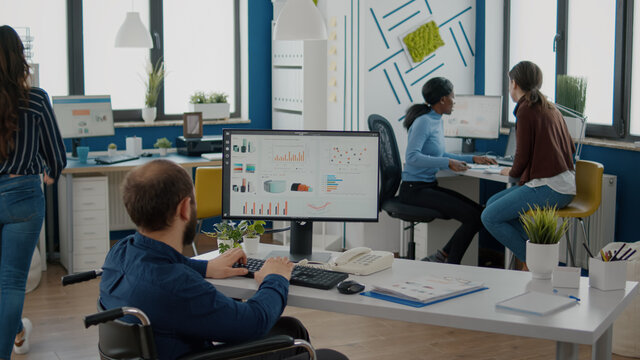 Paralized employee talking with african team leader woman sharing information pointing on graphs from computer. Colleague giving feedback discussing solution for project deadline in strat up office